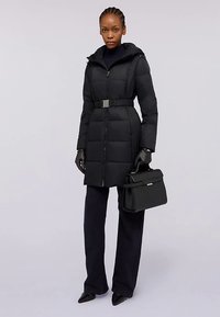 Svart vadderad jacka med huva och skärp i midjan. Har en dragkedja fram, strukturerat tyg och är kombinerad med svarta handskar och en handväska.