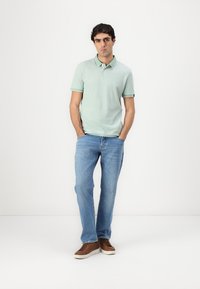Lys grøn polo skjorte med korte ærmer og krave, kombinert med blå denim jeans og brune sko. Stoffet ser mykt og pustende ut.