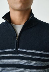 Marinefarbenes Zip-Up-Pullover mit geripptem Kragen und Streifen in Hellblau. Verfügt über einen goldenen Reißverschluss und eine weiche Textur.