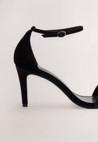 Crna sandala od brušene kože s visokim petama, tankom narukvicom oko gležnja s kopčom, otvorenim vrhom i suženom petom na neutralnoj pozadini.
