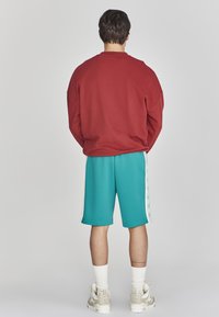Felpa rossa oversize, pantaloni corti turchesi con strisce bianche ai lati, calzini bianchi e sneakers di colore chiaro. Dettagli di cucitura visibili.