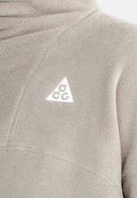 Pullover in fleece di un beige chiaro con colletto alto e un patch triangolare riflettente. Presenta un design senza cuciture e un tessuto testurizzato.