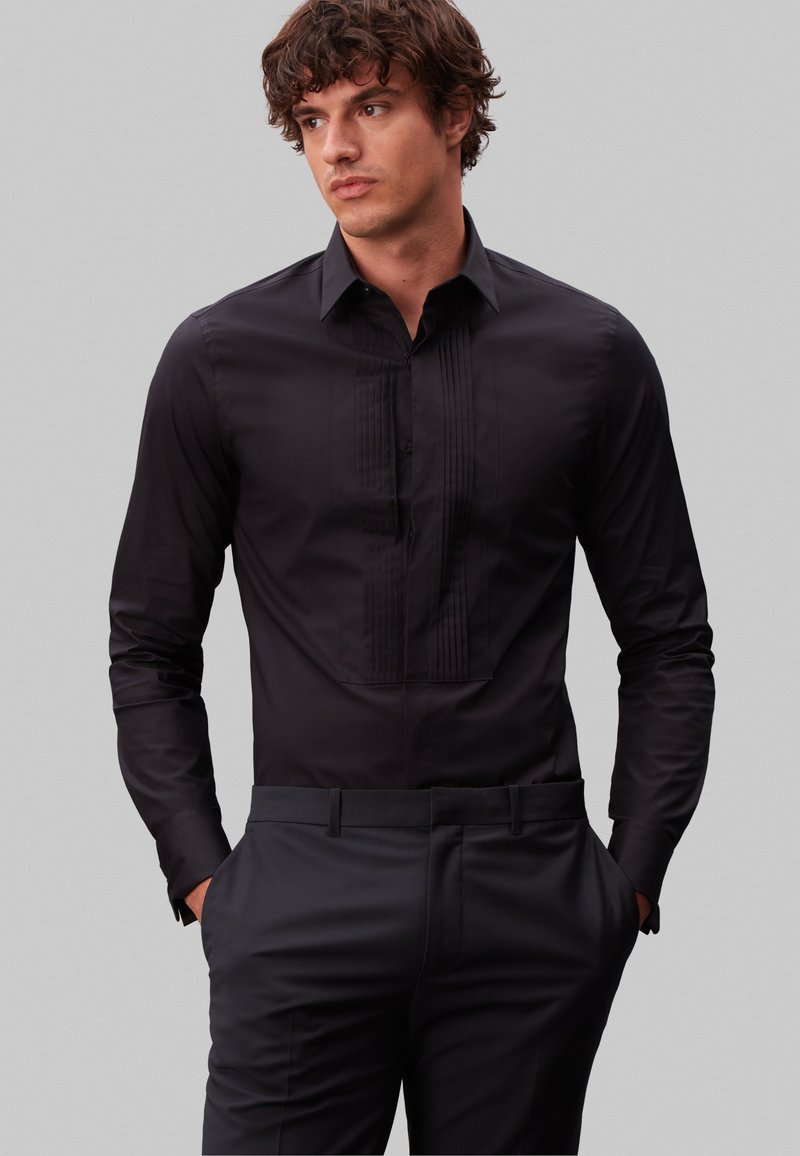 Homme aux cheveux bouclés portant une chemise noire cintrée avec un devant plissé et un pantalon noir, mains dans les poches, regardant légèrement sur le côté.