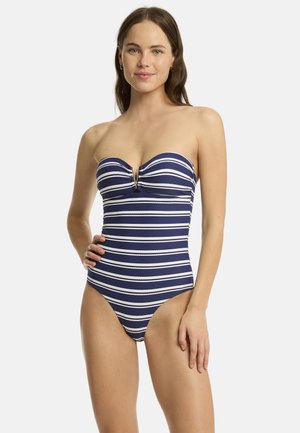 Kobieta w granatowo-biały, paskowany, jednoczęściowy strój kąpielowy bez ramiączek z metalowym akcentem w kształcie litery V na dekolcie, stoi z ręką na biodrze.