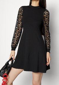 Robe noire avec un corsage ajusté et une jupe évasée. Les manches longues présentent une superposition en dentelle noire avec un motif festonné. Col haut simple.