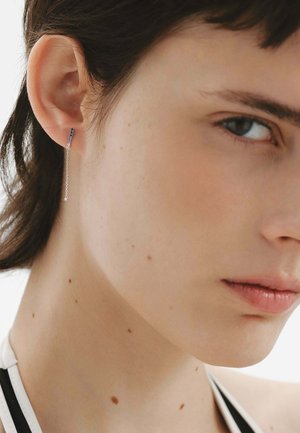Pendientes colgantes de plata con un acento de piedra preciosa azul, que presentan un diseño elegante y minimalista y una fina cadena que se extiende desde el lóbulo de la oreja.