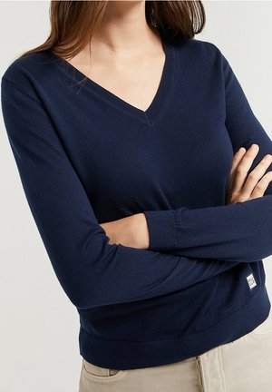Femme aux longs cheveux bruns portant un pull bleu marine à col en V, bras croisés, et un pantalon beige devant un fond clair uni.