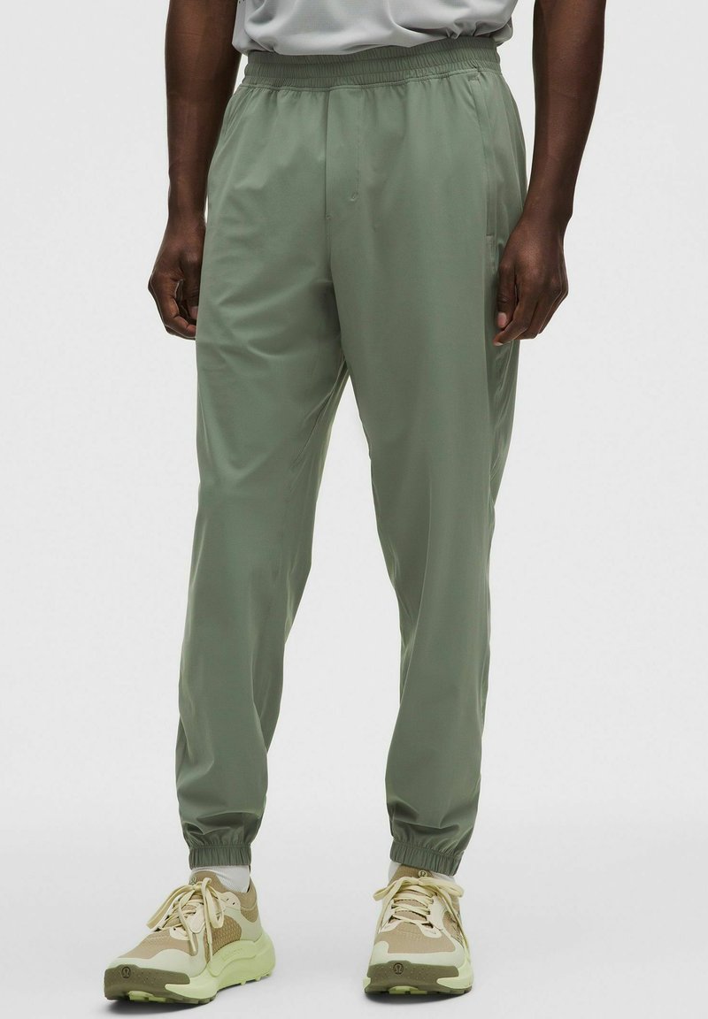 Uomo che indossa pantaloni jogger con coulisse elastiche color verde chiaro e sneakers beige, in piedi con le mani rilassate lungo i fianchi su sfondo neutro.