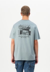 Férfi világoskék pólót visel, amelyen fekete grafikával egy tejbár boltja, egy szörfdeszka és a "Rip Curl Australia" felirat látható a hátsó részen.