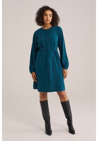 Tealfärgad klänning med rund halsringning, r gathered fabric vid axlarna och ett matchande bälte. Bärs med svarta knähöga stövlar. Slät textur.