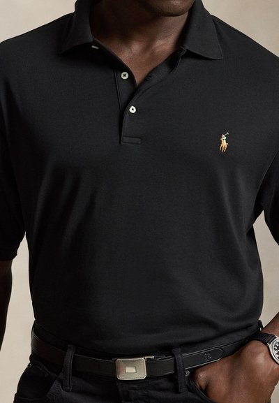 Mann trägt ein schwarzes Polohemd mit kleinem gesticktem Pferd-und-Reiter-Logo, schwarzen Gürtel und Armbanduhr, Hand in der Tasche.