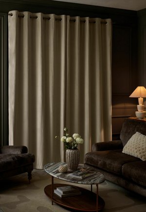 Wohnzimmer mit beigen Vorhängen, dunkelbraunem Samtsofa und Sessel, Marmor-Couchtisch mit Vase mit weißen Blumen und eingeschalteter Tischlampe.