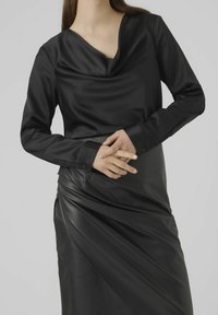 Haut noir à manches longues avec un col drapé, superposé sur une jupe en cuir noir à design drapé et texture lisse.