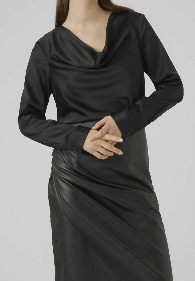 Haut noir à manches longues avec un col drapé, superposé sur une jupe en cuir noir à design drapé et texture lisse.