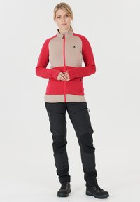 Röd och beige zip-jacka med texturerad tyg och tumhål, i kombination med svarta byxor med fickor med dragkedja och förstärkta knän.