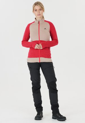 Rote und beige Zip-Jacke aus strukturiertem Material mit Daumenlöchern, kombiniert mit schwarzen Hosen mit Reißverschlusstaschen und verstärkten Knien.
