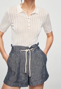 Žena v bílém polokošili s krátkým rukávem a vertikálními pruhy, spolu s modrými šortkami s vysokým pasem a bílým lanovým páskem.