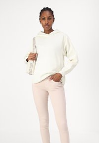 Sudadera color beige claro con capucha y cremalleras laterales, combinada con jeans rosa pálido. Textura suave y corte relajado. Bolso cruzado con correa de logo.
