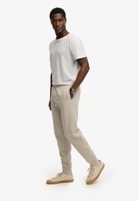 Hellgraues T-Shirt, leger sitzende beige Jogginghosen und hellfarbene Sneakers. Armbanduhr an der linken Hand; Modell steht im Profil vor einem schlichten Hintergrund.