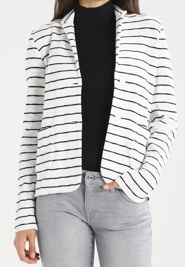 Blazer rayé blanc avec des rayures noires horizontales, col cranté, deux poches avant et une coupe décontractée, associé à un haut noir et un pantalon gris.