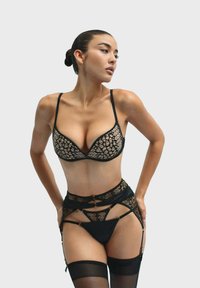 Zwarte kant bralette met beugels, voorzien van een geometrisch patroon. Bijpassende zwarte kousenband met uitsparingen en gouden accenten. Transparante kousen.