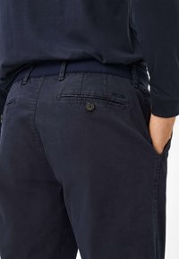 Marineblauwe katoenen broek met een gladde textuur, voorzien van twee achterzakken met knopen en een subtiele merketiket op de tailleband.