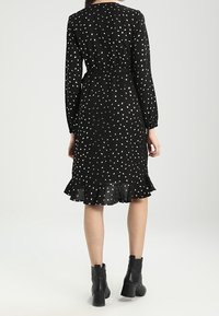 Robe noire à pois blancs, manches longues, ourlet à volants et taille cintrée. Portée avec des bottines noires. Texture de tissu lisse.