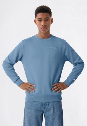 Ung mand med kort krøllet hår iført en lyseblå Champion sweatshirt og blå jeans, står med hænderne på hoften mod hvid baggrund.