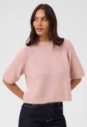 Femme aux longs cheveux bruns portant un pull court doux et duveteux rose pâle et un jean bleu foncé, debout les mains derrière le dos.