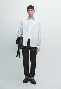 Camicia bianca a maniche lunghe sopra una T-shirt bianca, abbinata a jeans scuri. Il modello tiene una borsa in pelle nera e indossa scarpe scure con una finitura lucida.