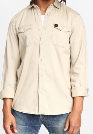Beige overhemd met lange mouwen, gemaakt van zachte stof, met een knoopsluiting aan de voorkant, twee borstzakken en opgerolde mouwen.