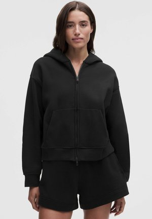 Vrouw met halflang donker haar draagt een zwarte hoodie met rits en bijpassende zwarte shorts, staand tegen een effen lichte achtergrond.