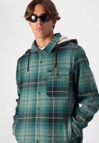 Rutig flanelljacka i teal, svart och grått. Har en huva, bröstficka och knäppningar. Texturerat tyg med ett rutmönster.