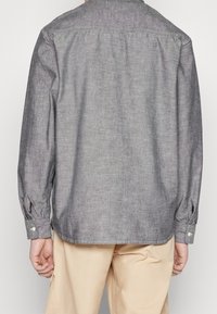 Achteraanzicht van een persoon die een grijs overhemd met lange mouwen en beige broek draagt, tegen een effen achtergrond.