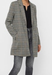 Šedý kostkovaný blazer se strukturovaným designem, jednobarevný přední díl a dvě boční kapsy. Kombinováno s černým topem a šedými džíny.