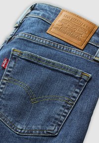 Jean en denim bleu plié pour montrer la poche arrière avec des coutures jaunes, une étiquette rouge Levi's et un patch en cuir beige de la marque Levi Strauss & Co.