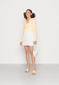 Top amarelo em peplum com laço à frente, vestido branco de renda, mules amarelas de salto e uma bolsa branca com alça texturizada.