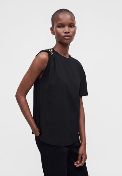 Femme aux cheveux courts portant un haut noir sans manches avec un détail doré à l'épaule et un pantalon noir, debout avec une main dans la poche.
