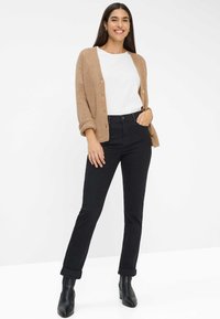 Cardigan marrone chiaro su una camicia bianca, abbinato a jeans neri a vita alta arrotolati alla caviglia, accentuato da stivali neri con tacco.