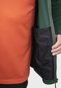 Schichtkleidung besteht aus einem orangefarbenen strukturierten Hemd unter einer grünen Fleecejacke mit einer schwarzen Innentasche und einem Reißverschluss.