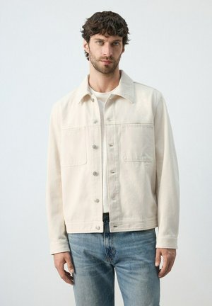 Hombre de cabello oscuro y barba que lleva una chaqueta abotonada de color beige claro sobre una camisa blanca y vaqueros azules, de pie frente a un fondo sencillo.