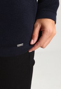 Maglione a coste blu navy con superficie testurizzata. Presenta una piccola etichetta in metallo argento sull'orlo. Una mano poggiata sul lato del maglione. Pantaloni neri visibili.