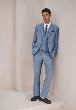 Ung mand i blå tre-delt jakkesæt, hvid skjorte, mønstret sort slips, sorte sko, stående med hænderne i lommerne mod en beige væg.