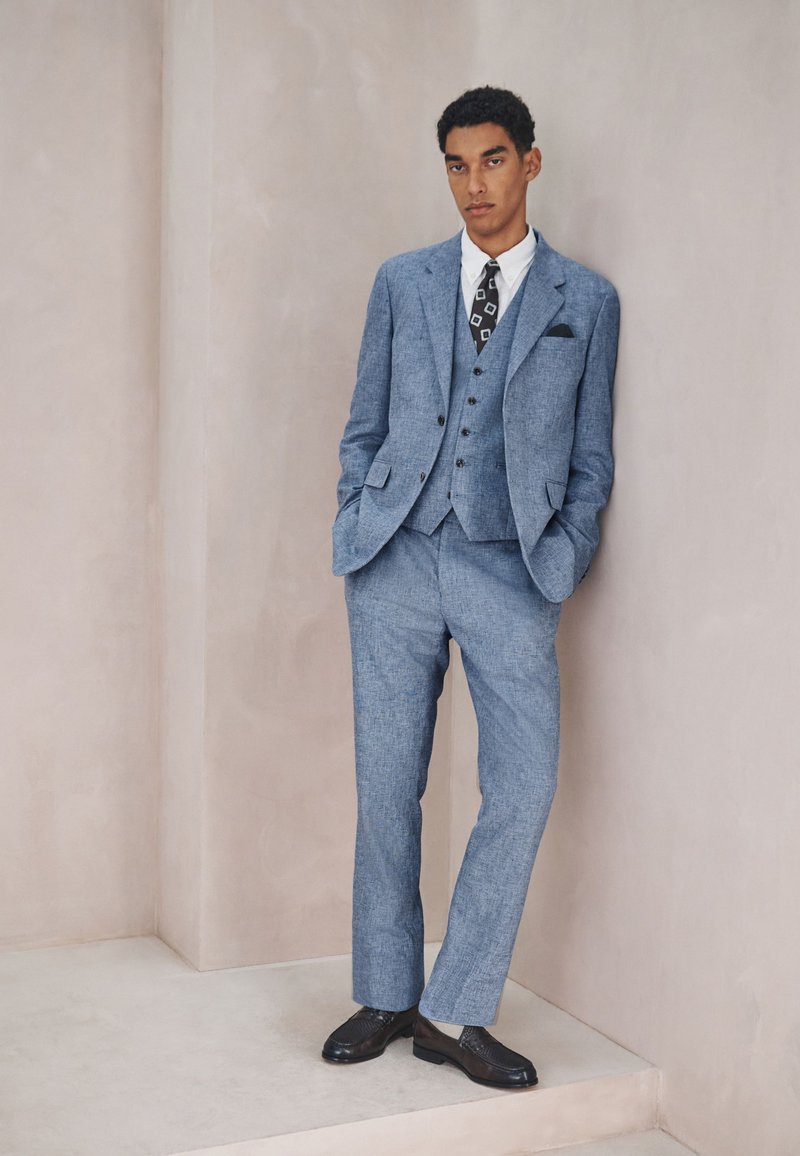 Jeune homme en costume trois pièces bleu, chemise blanche, cravate noire à motifs, chaussures noires, debout les mains dans les poches contre un mur beige.