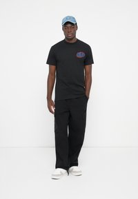 Sort kortærmet T-shirt med rød og blå "HUF" logo, parret med løstsiddende sorte cargo bukser og hvide sneakers. Lyseblå kasket.