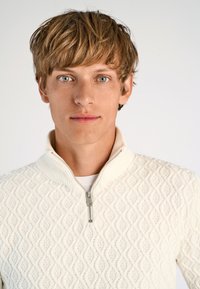 Witte gebreide trui met een rits bij de staande kraag, met een gemêleerd ruitpatroon. Model heeft kort, tousled haar en blauwe ogen.