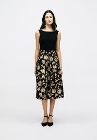 Μαύρο αμάνικο τοπ σε συνδυασμό με φούστα σε μήκος έως το γόνατο, με χρυσό ανθικό μοτίβο. Αξεσουάρ περιλαμβάνουν μαύρα παπούτσια με λουράκι στον αστράγαλο.