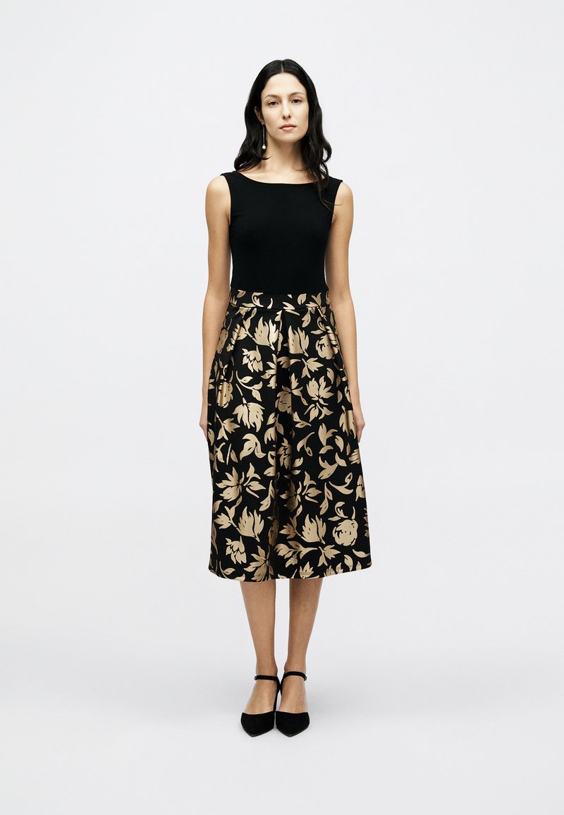 Μαύρο αμάνικο τοπ σε συνδυασμό με φούστα σε μήκος έως το γόνατο, με χρυσό ανθικό μοτίβο. Αξεσουάρ περιλαμβάνουν μαύρα παπούτσια με λουράκι στον αστράγαλο.