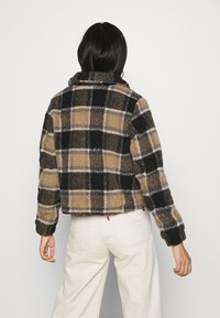 Persona con cabello negro que lleva una chaqueta de forro polar corta de cuadros en negro, beige y gris y pantalones anchos color blanco roto, mirando hacia el lado opuesto.