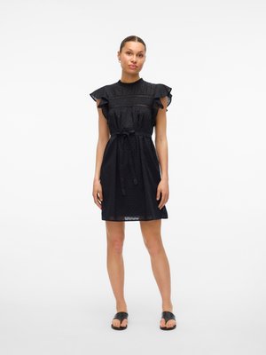 Femme debout portant une courte robe noire à manches volantées et ceinture à la taille, chaussée de sandales noires, sur un fond blanc uni.